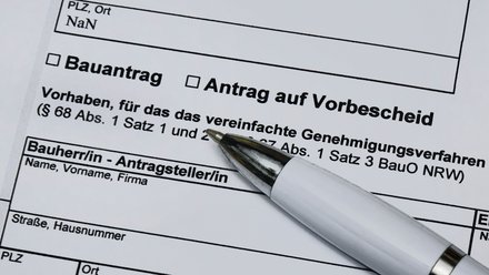 Ein stark vergrößerter Ausschnitt eines offiziellen Formulars zeigt die Felder „Bauantrag“ und „Antrag auf Vorbescheid“ sowie Textpassagen zu einem vereinfachten Genehmigungsverfahren nach der Bauordnung, ein weißer Kugelschreiber liegt schräg auf dem Papier und weist auf den auszufüllenden Bereich, wodurch das Bild administrative Abläufe rund um Planen und Bauen sichtbar macht und für die formalen, rechtlichen Rahmenbedingungen steht, die Baukultur und bauliche Umsetzung begleiten.