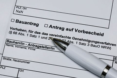 Ein stark vergrößerter Ausschnitt eines offiziellen Formulars zeigt die Felder „Bauantrag“ und „Antrag auf Vorbescheid“ sowie Textpassagen zu einem vereinfachten Genehmigungsverfahren nach der Bauordnung, ein weißer Kugelschreiber liegt schräg auf dem Papier und weist auf den auszufüllenden Bereich, wodurch das Bild administrative Abläufe rund um Planen und Bauen sichtbar macht und für die formalen, rechtlichen Rahmenbedingungen steht, die Baukultur und bauliche Umsetzung begleiten.