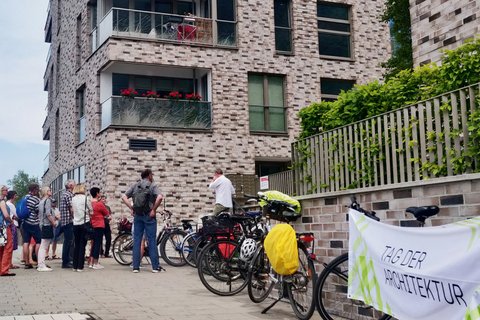 Gruppe von Menschen steht vor einem mehrstöckigen Wohngebäude aus roten Backsteinen mit mehreren Balkonen und großen Fenstern. Rechts im Bild Fahrräder und ein Plakat mit Text 'ZUBER ARCHITEKTUR'. Eine Efeu-bewachsene grüne Zaunkonstruktion ist sichtbar. Ein Ort von architektonischem Interesse, möglicherweise eine Architekturführung oder Besichtigung."