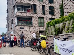 Gruppe von Menschen steht vor einem mehrstöckigen Wohngebäude aus roten Backsteinen mit mehreren Balkonen und großen Fenstern. Rechts im Bild Fahrräder und ein Plakat mit Text 'ZUBER ARCHITEKTUR'. Eine Efeu-bewachsene grüne Zaunkonstruktion ist sichtbar. Ein Ort von architektonischem Interesse, möglicherweise eine Architekturführung oder Besichtigung."
