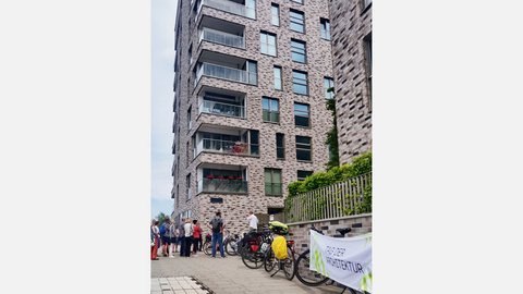 Gruppe von Menschen steht vor einem mehrstöckigen Wohngebäude aus roten Backsteinen mit mehreren Balkonen und großen Fenstern. Rechts im Bild Fahrräder und ein Plakat mit Text 'ZUBER ARCHITEKTUR'. Eine Efeu-bewachsene grüne Zaunkonstruktion ist sichtbar. Ein Ort von architektonischem Interesse, möglicherweise eine Architekturführung oder Besichtigung."