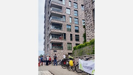 Gruppe von Menschen steht vor einem mehrstöckigen Wohngebäude aus roten Backsteinen mit mehreren Balkonen und großen Fenstern. Rechts im Bild Fahrräder und ein Plakat mit Text 'ZUBER ARCHITEKTUR'. Eine Efeu-bewachsene grüne Zaunkonstruktion ist sichtbar. Ein Ort von architektonischem Interesse, möglicherweise eine Architekturführung oder Besichtigung."