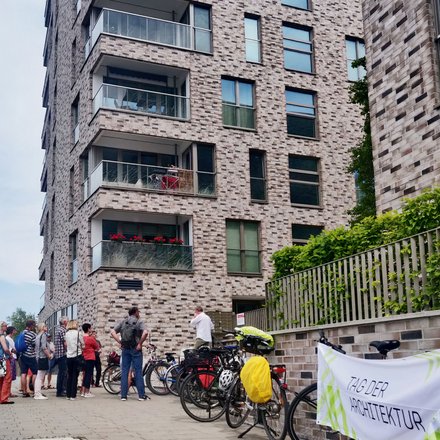Gruppe von Menschen steht vor einem mehrstöckigen Wohngebäude aus roten Backsteinen mit mehreren Balkonen und großen Fenstern. Rechts im Bild Fahrräder und ein Plakat mit Text 'ZUBER ARCHITEKTUR'. Eine Efeu-bewachsene grüne Zaunkonstruktion ist sichtbar. Ein Ort von architektonischem Interesse, möglicherweise eine Architekturführung oder Besichtigung."