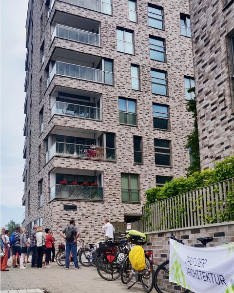 Gruppe von Menschen steht vor einem mehrstöckigen Wohngebäude aus roten Backsteinen mit mehreren Balkonen und großen Fenstern. Rechts im Bild Fahrräder und ein Plakat mit Text 'ZUBER ARCHITEKTUR'. Eine Efeu-bewachsene grüne Zaunkonstruktion ist sichtbar. Ein Ort von architektonischem Interesse, möglicherweise eine Architekturführung oder Besichtigung."