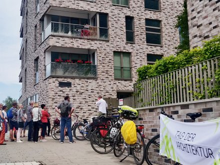 Gruppe von Menschen steht vor einem mehrstöckigen Wohngebäude aus roten Backsteinen mit mehreren Balkonen und großen Fenstern. Rechts im Bild Fahrräder und ein Plakat mit Text 'ZUBER ARCHITEKTUR'. Eine Efeu-bewachsene grüne Zaunkonstruktion ist sichtbar. Ein Ort von architektonischem Interesse, möglicherweise eine Architekturführung oder Besichtigung."