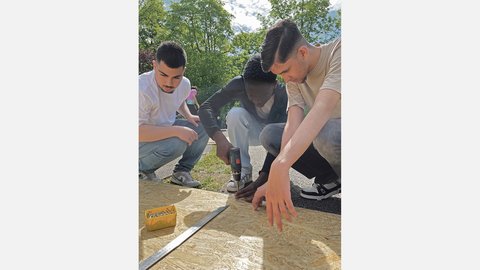 "Drei junge Männer arbeiten gemeinsam im Freien an einer großen Holzplatte. Einer von ihnen kniet und schraubt mit einem Akkuschrauber eine Schraube in das Holz, während die beiden anderen aufmerksam zusehen. Auf der Platte liegen ein Winkelmaß und eine gelbe Box mit Schrauben."