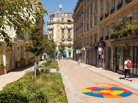 "Breite fußgängerfreundliche Straße in Paris mit historischen Sandsteinfassaden rechts und links. Links befindet sich ein breiter Grünstreifen mit Bäumen und Grün in Pflanzgefäßen. Rechts sind Geschäftsschaufenster mit Rolladen. Menschen gehen spazieren. Ein farbiger kreisförmiger Kunstmotiv ist auf dem Belag aufgebracht. Eine begrünte, autofreie Straße."