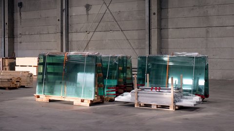  Gestapelte Glasscheiben auf Paletten, in Folie verpackt, in einer Industriehalle; daneben Holz- und Metallteile.
