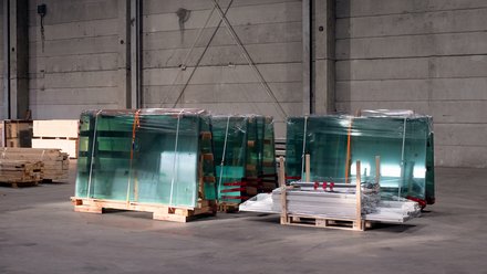  Gestapelte Glasscheiben auf Paletten, in Folie verpackt, in einer Industriehalle; daneben Holz- und Metallteile.