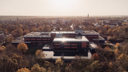Luftaufnahme eines weitläufigen Schulkomplexes mit Flachdächern und orangefarbenen Fassadenbändern, umgeben von herbstlichen Bäumen; Sonnenspiegelung auf dem Dach, Stadtpanorama mit Kirchturm im Hintergrund.