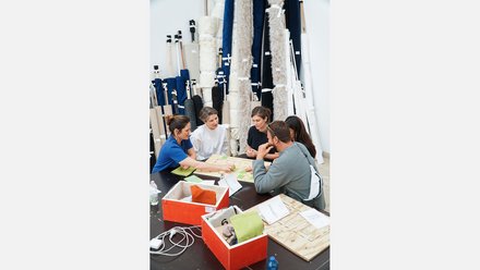 Eine Gruppe von fünf Personen sitzt konzentriert um einen Tisch und arbeitet an einem Modell oder Brettspiel namens „CIRCUIT“. Im Hintergrund lehnen zahlreiche hohe Stoffrollen in verschiedenen Farben und Texturen an der Wand.