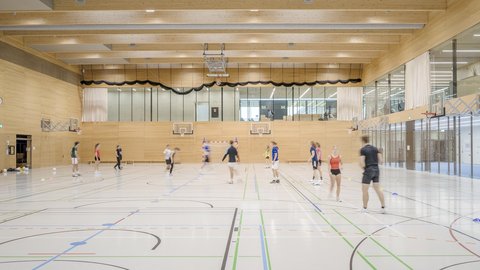 Helle Dreifachsporthalle mit Holzdecke und Personen beim Training.