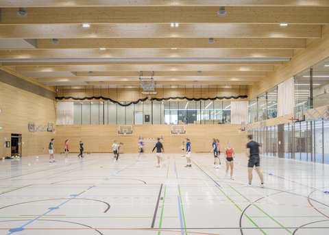 Helle Dreifachsporthalle mit Holzdecke und Personen beim Training.