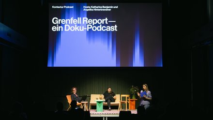 Bühnensituation einer Veranstaltung des kntxtr-Podcasts. Eine riesige Leinwand zeigt den Titel „Grenfell Report — ein Doku-Podcast“. Auf der Bühne sitzen zwei Frauen auf einem beigen Sofa und eine dritte Frau steht rechts daneben und moderiert mit einer Mappe in der Hand vor Publikum.