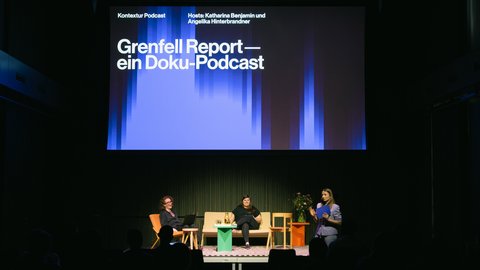 Bühnensituation einer Veranstaltung des kntxtr-Podcasts. Eine riesige Leinwand zeigt den Titel „Grenfell Report — ein Doku-Podcast“. Auf der Bühne sitzen zwei Frauen auf einem beigen Sofa und eine dritte Frau steht rechts daneben und moderiert mit einer Mappe in der Hand vor Publikum.