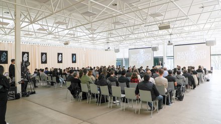 Großer Vortragssaal mit Publikum auf Stuhlreihen, das einem Vortrag auf zwei Projektionsleinwänden folgt.