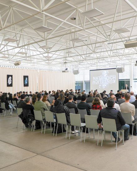 Großer Vortragssaal mit Publikum auf Stuhlreihen, das einem Vortrag auf zwei Projektionsleinwänden folgt.