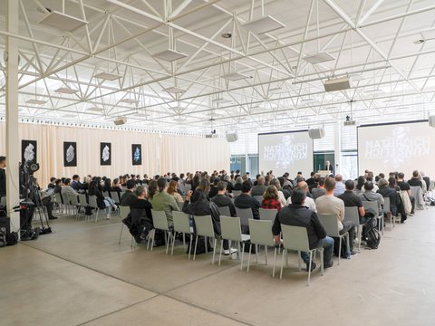 Großer Vortragssaal mit Publikum auf Stuhlreihen, das einem Vortrag auf zwei Projektionsleinwänden folgt.
