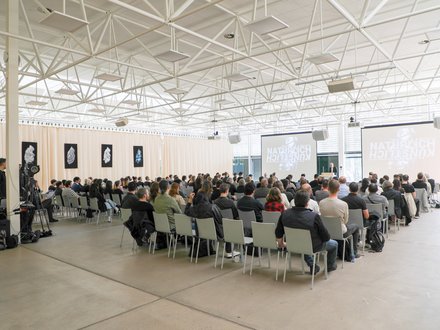 Großer Vortragssaal mit Publikum auf Stuhlreihen, das einem Vortrag auf zwei Projektionsleinwänden folgt.