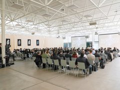 Großer Vortragssaal mit Publikum auf Stuhlreihen, das einem Vortrag auf zwei Projektionsleinwänden folgt.