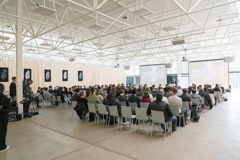 Großer Vortragssaal mit Publikum auf Stuhlreihen, das einem Vortrag auf zwei Projektionsleinwänden folgt.