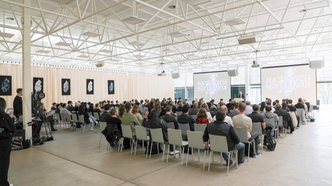Großer Vortragssaal mit Publikum auf Stuhlreihen, das einem Vortrag auf zwei Projektionsleinwänden folgt.