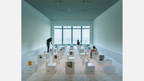 Alt-Text: "Moderner Ausstellungsraum mit hellblaugrüner Wandfarbe und graugrüner Decke. Ein großes Fenster mit dunklen Streben bietet Ausblick auf eine Stadt-Skyline. Der Raum ist mit zahlreichen weißen kubischen Ausstellungsständern gefüllt, die verschiedene Projekte präsentieren. Drei Besucher bewegen sich durch den Raum und betrachten die Exponate. Der Boden ist hell und glatt."