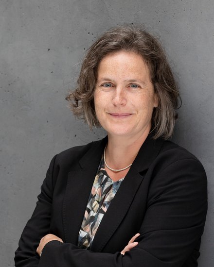 Porträtfoto: Person mit kurzem, leicht gewelltem Haar, lächelt, trägt schwarzen Blazer, gemustertes Oberteil und feine Kette; vor grauer Betonwand, Arme verschränkt.