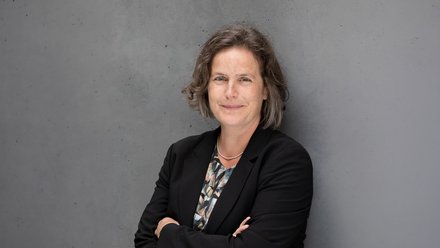 Porträtfoto: Person mit kurzem, leicht gewelltem Haar, lächelt, trägt schwarzen Blazer, gemustertes Oberteil und feine Kette; vor grauer Betonwand, Arme verschränkt.