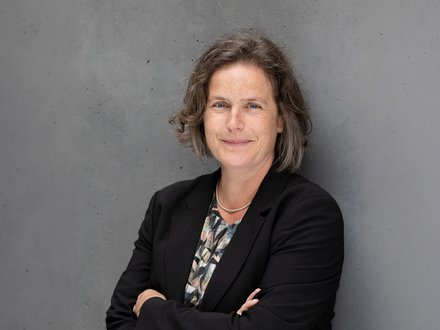 Porträtfoto: Person mit kurzem, leicht gewelltem Haar, lächelt, trägt schwarzen Blazer, gemustertes Oberteil und feine Kette; vor grauer Betonwand, Arme verschränkt.