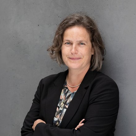 Porträtfoto: Person mit kurzem, leicht gewelltem Haar, lächelt, trägt schwarzen Blazer, gemustertes Oberteil und feine Kette; vor grauer Betonwand, Arme verschränkt.