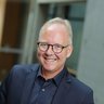 Ein Porträt zeigt eine einzelne erwachsene Person mit Brille vor unscharfem architektonischem Hintergrund, frontal zur Kamera ausgerichtet und freundlich lächelnd, wodurch das Bild eine professionelle Darstellung vermittelt und für fachliche Expertise, Engagement und persönliche Verantwortung innerhalb eines architektonischen oder kulturellen Kontextes steht.