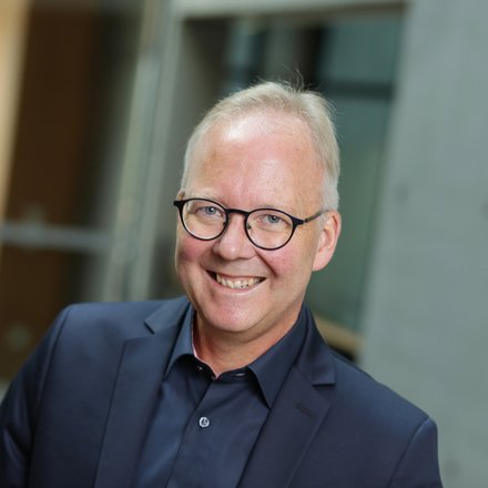 Ein Porträt zeigt eine einzelne erwachsene Person mit Brille vor unscharfem architektonischem Hintergrund, frontal zur Kamera ausgerichtet und freundlich lächelnd, wodurch das Bild eine professionelle Darstellung vermittelt und für fachliche Expertise, Engagement und persönliche Verantwortung innerhalb eines architektonischen oder kulturellen Kontextes steht.