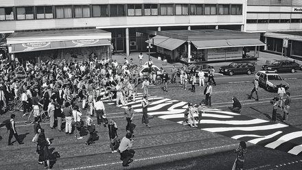 Eine historische Schwarz-Weiß-Fotografie aus der Vogelperspektive. Eine Menschenmenge überquert eine Straße, auf die ein großflächiges, grafisches Muster (ähnlich einem Zebra-Look) gemalt wurde.