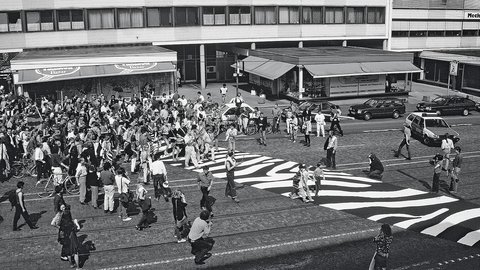 Eine historische Schwarz-Weiß-Fotografie aus der Vogelperspektive. Eine Menschenmenge überquert eine Straße, auf die ein großflächiges, grafisches Muster (ähnlich einem Zebra-Look) gemalt wurde.