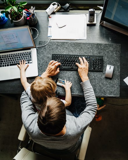 Arbeitsumfeld mit Kind: Ein Elternteil arbeitet am Computer, während ein Kind auf dem Tisch sitzt und mit einem Smartphone spielt.