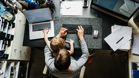 Arbeitsumfeld mit Kind: Ein Elternteil arbeitet am Computer, während ein Kind auf dem Tisch sitzt und mit einem Smartphone spielt.