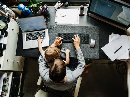 Arbeitsumfeld mit Kind: Ein Elternteil arbeitet am Computer, während ein Kind auf dem Tisch sitzt und mit einem Smartphone spielt.