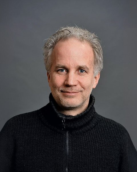 Porträtfoto eines Mannes mittleren Alters mit ergrauten, lockigen Haaren und Bart. Er trägt einen schwarzen Pullover mit Reißverschluss über einem strukturierten Unterhemd und blickt freundlich in die Kamera. Der Hintergrund ist neutral grau gehalten.