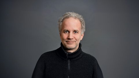 Porträtfoto eines Mannes mittleren Alters mit ergrauten, lockigen Haaren und Bart. Er trägt einen schwarzen Pullover mit Reißverschluss über einem strukturierten Unterhemd und blickt freundlich in die Kamera. Der Hintergrund ist neutral grau gehalten.