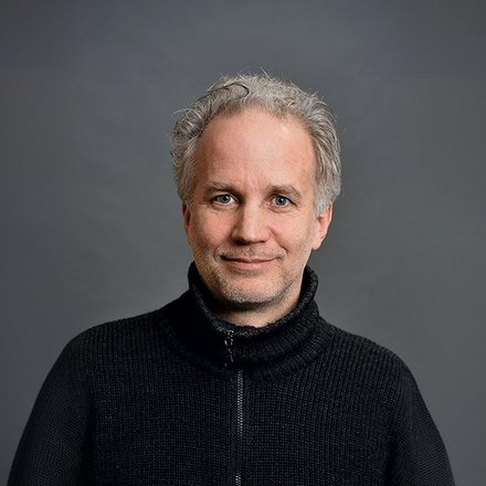Porträtfoto eines Mannes mittleren Alters mit ergrauten, lockigen Haaren und Bart. Er trägt einen schwarzen Pullover mit Reißverschluss über einem strukturierten Unterhemd und blickt freundlich in die Kamera. Der Hintergrund ist neutral grau gehalten.