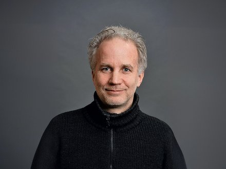 Porträtfoto eines Mannes mittleren Alters mit ergrauten, lockigen Haaren und Bart. Er trägt einen schwarzen Pullover mit Reißverschluss über einem strukturierten Unterhemd und blickt freundlich in die Kamera. Der Hintergrund ist neutral grau gehalten.