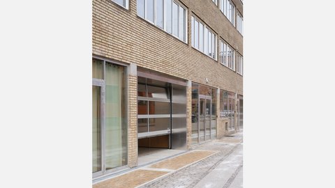 Außenansicht eines Gebäudes mit gelblicher Fassade. Im Erdgeschoss befinden sich große Fensterfronten; im Obergeschoss befinden sich gleichmäßige Fensterreihen