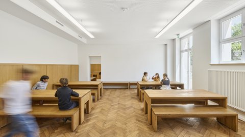 Heller Raum mit Holzparkett, Holztischen und -bänken, an denen mehrere Kinder sitzen und sich unterhalten