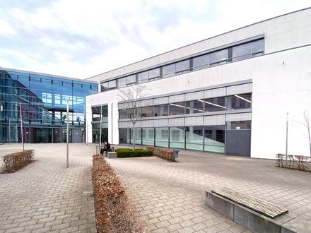 Moderner Hochschulcampus mit hellen Gebäudefassaden, großflächigen Glasfronten und einem gepflasterten Innenhof mit Sitzbänken und einzelnen Bäumen.