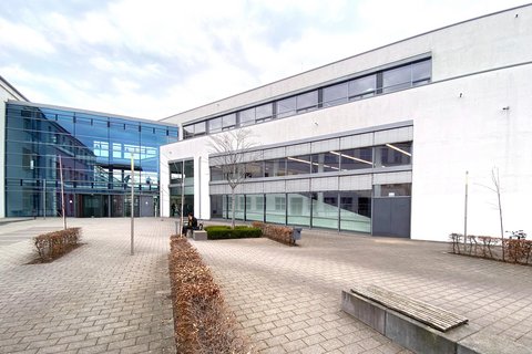 Moderner Hochschulcampus mit hellen Gebäudefassaden, großflächigen Glasfronten und einem gepflasterten Innenhof mit Sitzbänken und einzelnen Bäumen.