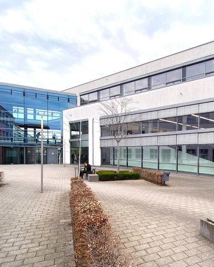 Moderner Hochschulcampus mit hellen Gebäudefassaden, großflächigen Glasfronten und einem gepflasterten Innenhof mit Sitzbänken und einzelnen Bäumen.