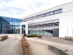 Moderner Hochschulcampus mit hellen Gebäudefassaden, großflächigen Glasfronten und einem gepflasterten Innenhof mit Sitzbänken und einzelnen Bäumen.