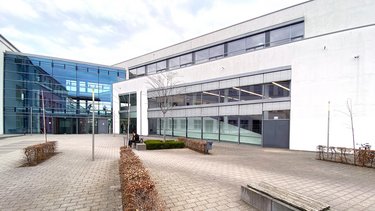 Moderner Hochschulcampus mit hellen Gebäudefassaden, großflächigen Glasfronten und einem gepflasterten Innenhof mit Sitzbänken und einzelnen Bäumen.
