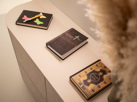 "Auf einem hellen beige-weißen zylindrischen Tisch arrangiert: Oben links ein schwarzes Notizbuch mit bunten Post-its (rosa, gelb, grün) und einen Stift in Vogel-Form. Mitte ein dunkelbraunes Buch mit weißem 'X'-Logo. Unten rechts ein antik wirkendes Buch mit goldenem ornamentalem Muster und dunklem Einband. Im unscharfen Hintergrund ein braun gefärbtes Objekt."